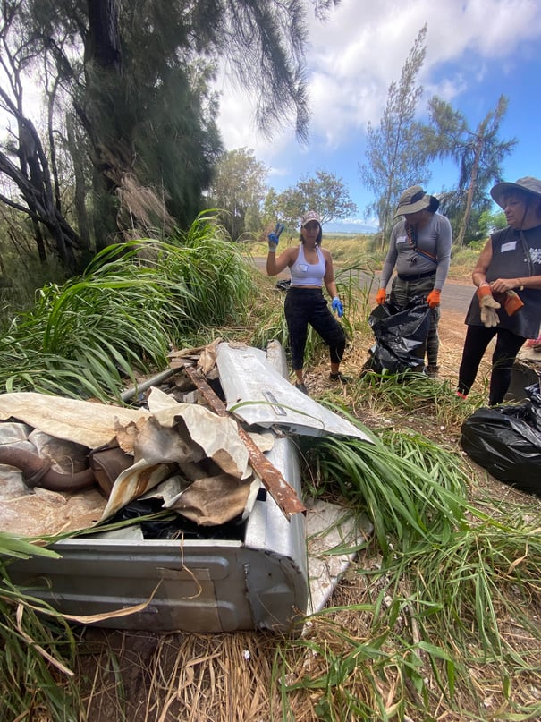 Volunteers removing metal and household debris from tall grass beside Holomua Road