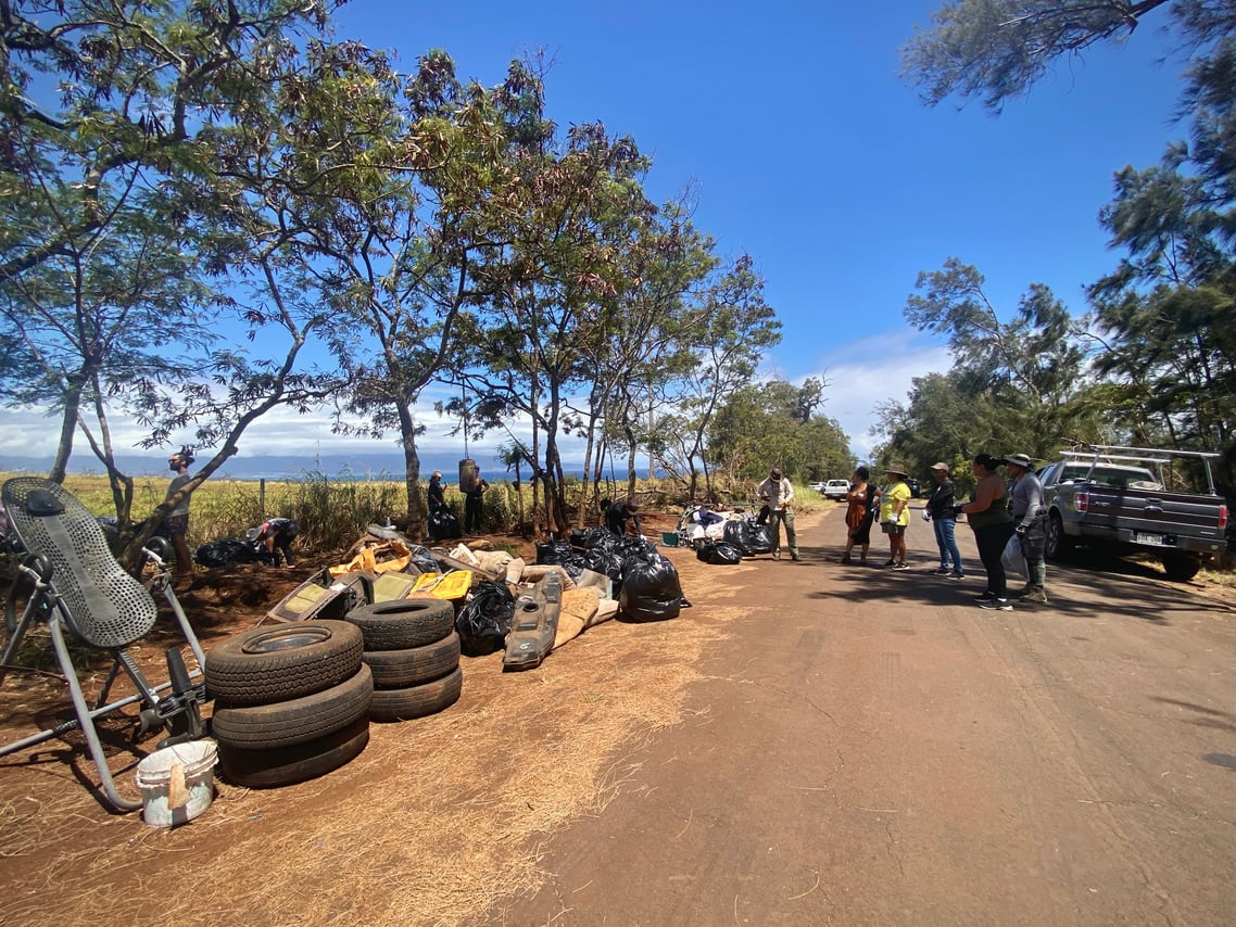 Tires, chairs, equipment, and bagged waste organized along Holomua Road for removal