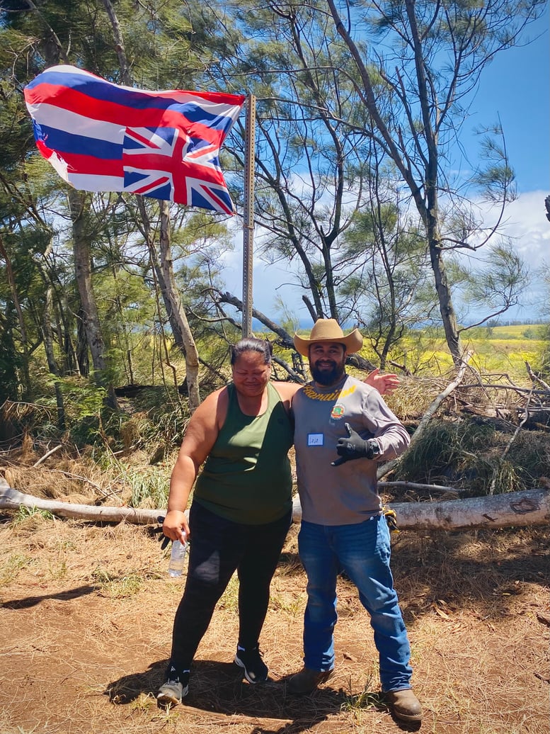 Two cleanup volunteers standing beneath the Hawaiʻi flag beside Holomua Road